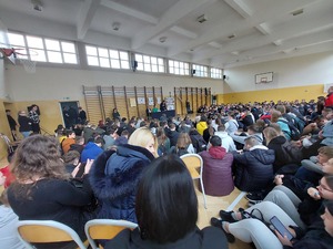 na zdjęciu widoczna siedząca na sali młodzież, w tle widoczne osoby siedzące przy stole, prowadzące konferencję oraz stojąca policjantka trzymająca mikrofon w ręce