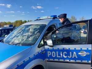 radiowóz policyjny, obok stojący policjant, a wewnątrz a kierownicą siedzi chłopiec