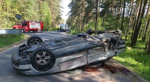 Zdjęcia ze zdarzenia drogowego w Wilczej Woli. Na pierwszym planie widać przewrócony na dach szary samochód, Z tyłu pojazdu widać biały samochód i znajdujących się obok ludzi oraz pojazd straży pożarnej.