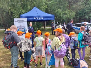 dzieci stojące przez namiotem z napisem POLICJA, w tle policjanta z mikrofonem