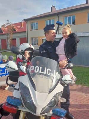 policjant trzymający dziecko na rękach, obok na motocyklu policyjnym siedzi dziecko